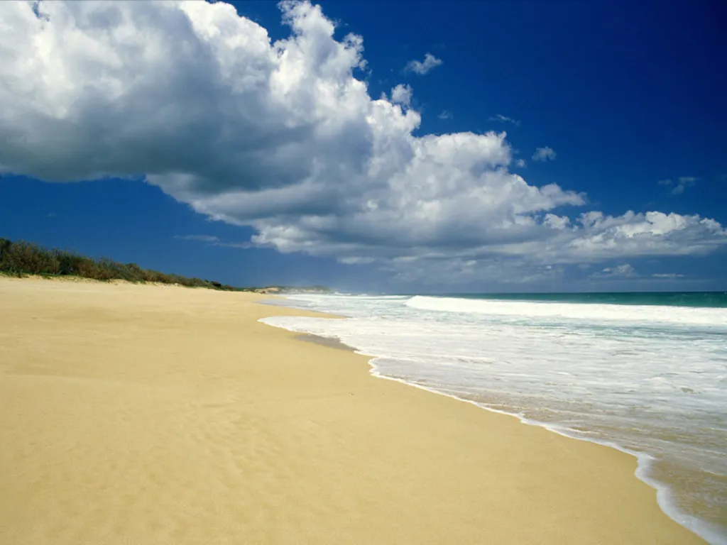 A wide sandy beach curves along the coast with gentle waves breaking on the shore under a bright blue sky with scattered clouds.