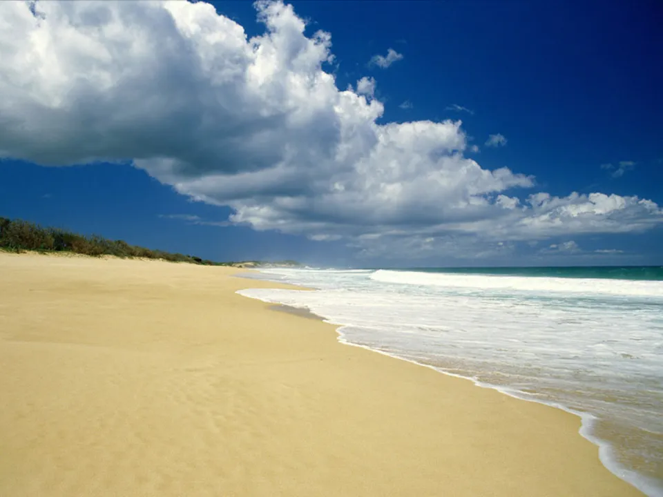 A wide sandy beach curves along the coast with gentle waves breaking on the shore under a bright blue sky with scattered clouds.