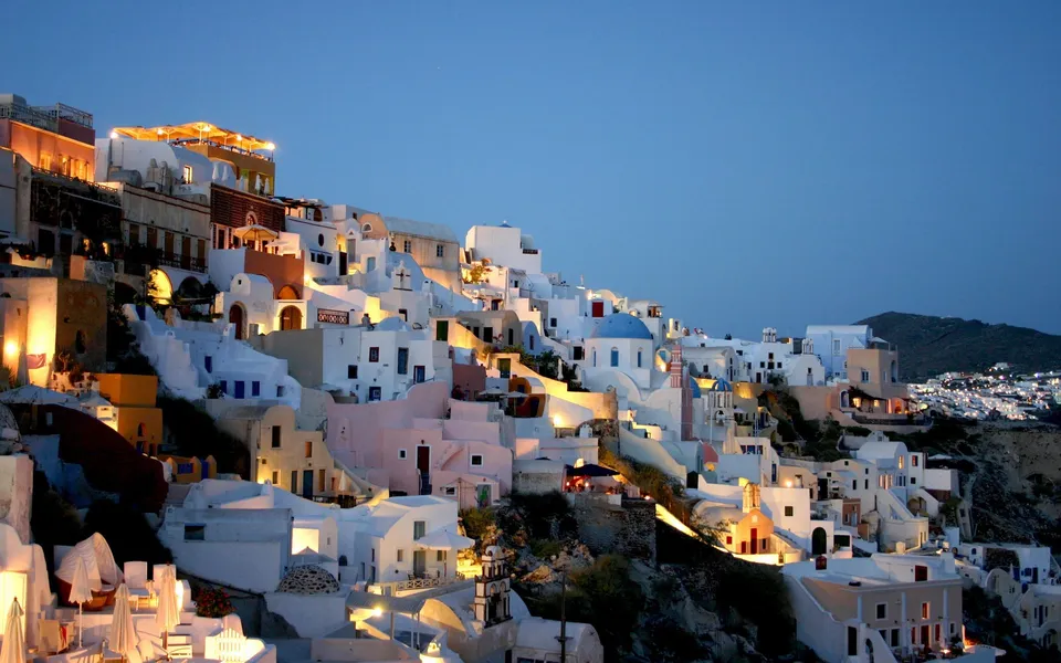 Whitewashed buildings with blue domes cascade down a hillside at dusk, illuminated by warm lights.