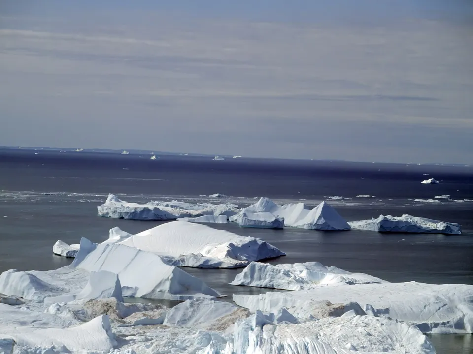 Numerous icebergs of various sizes are scattered across the dark ocean under a cloudy sky.