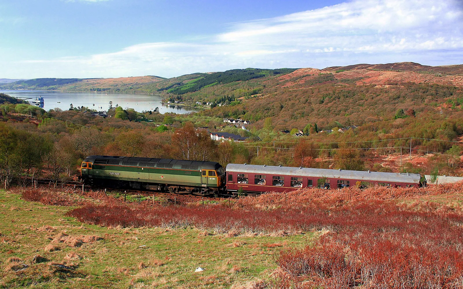 A green and maroon train travels along tracks through a landscape of hills, trees, and a distant loch.