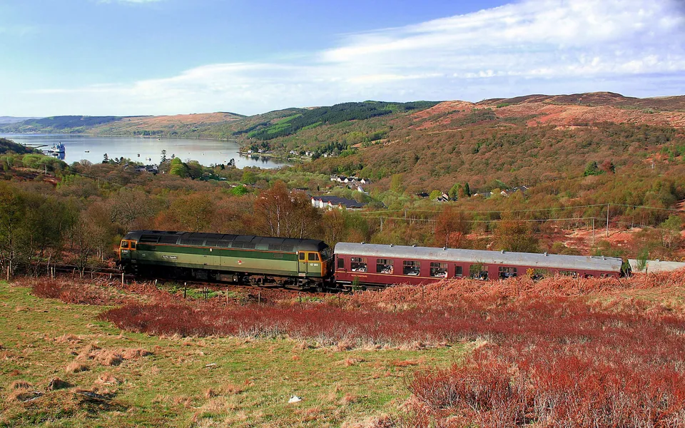 A green and maroon train travels along tracks through a landscape of hills, trees, and a distant loch.