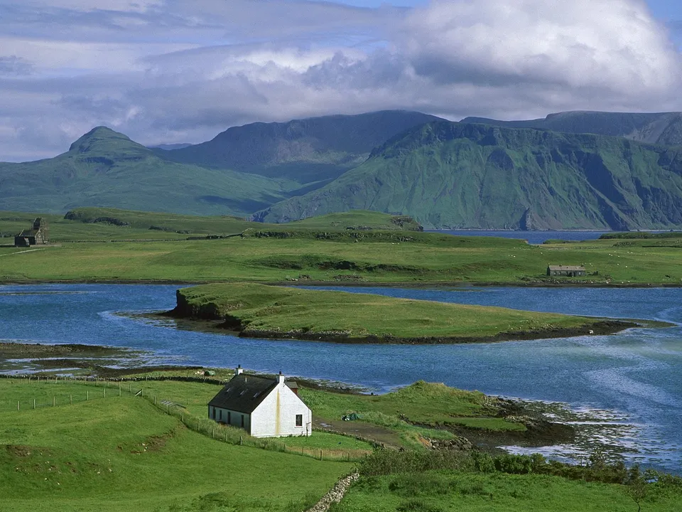 A white cottage sits on a green hill overlooking a blue bay with mountains in the background.