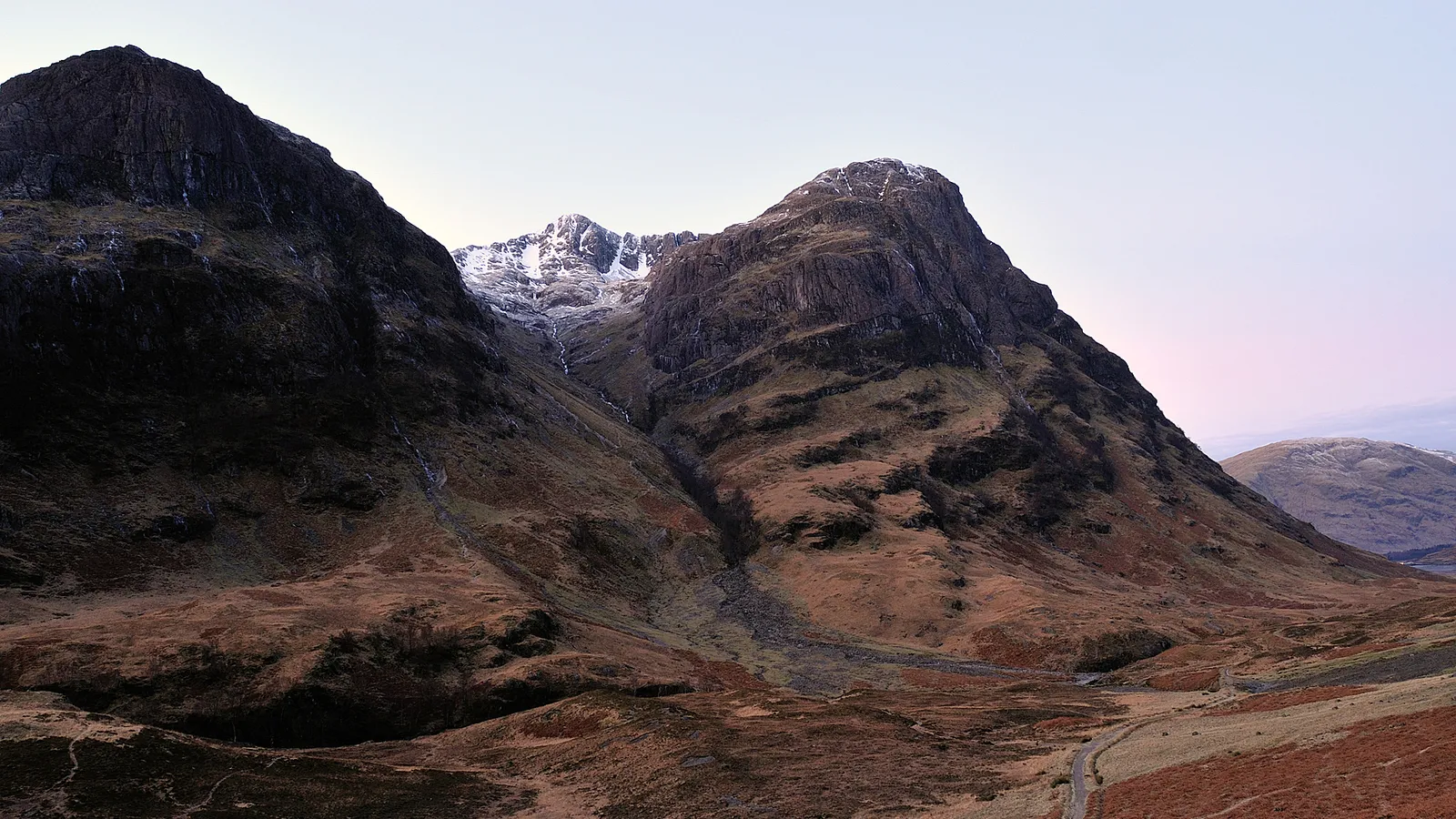 A wide shot of rugged, dark mountains with patches of snow on the peaks, under a pale sky.