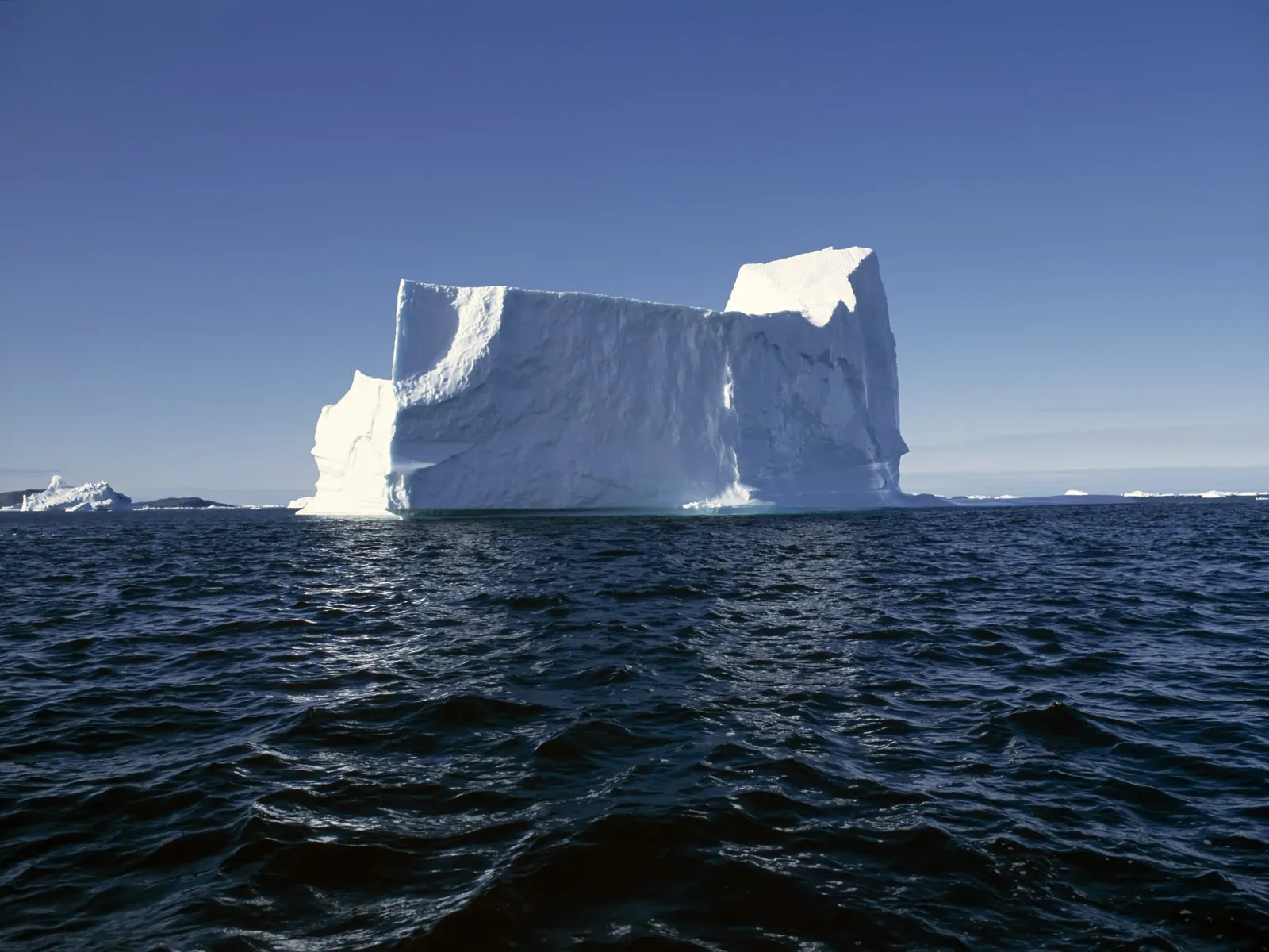 A colossal iceberg with a sculpted white surface floats in a deep blue, rippling ocean.