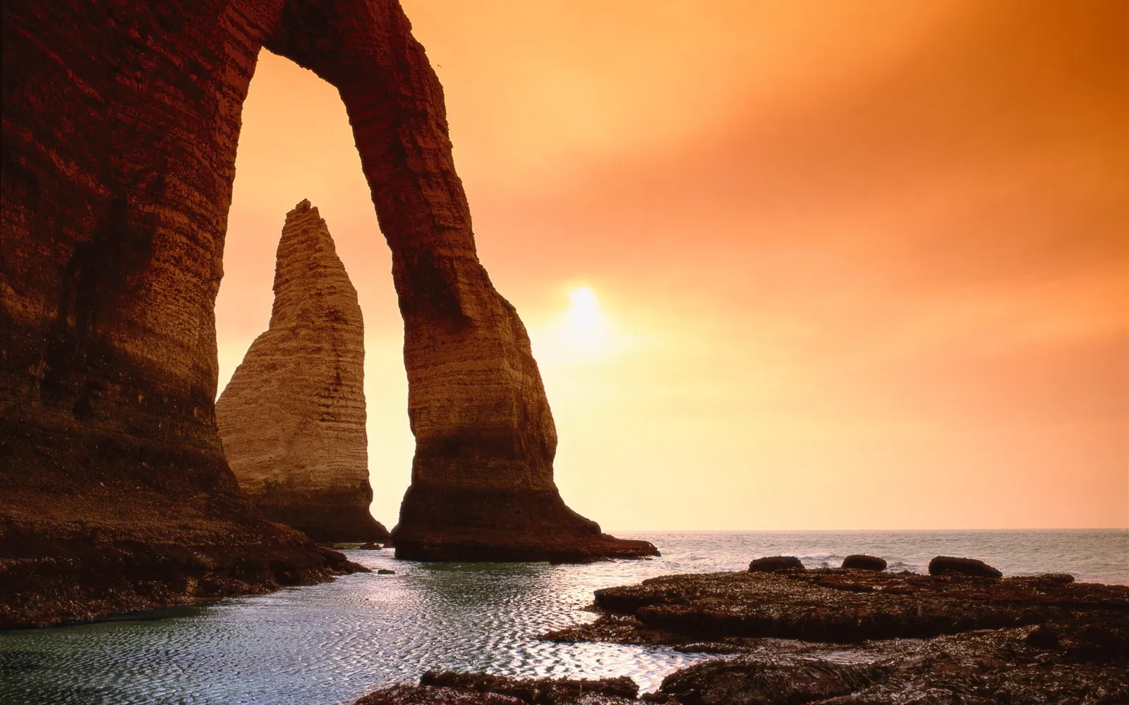 A natural rock arch frames a tall sea stack against a vibrant orange sunset sky over the ocean.