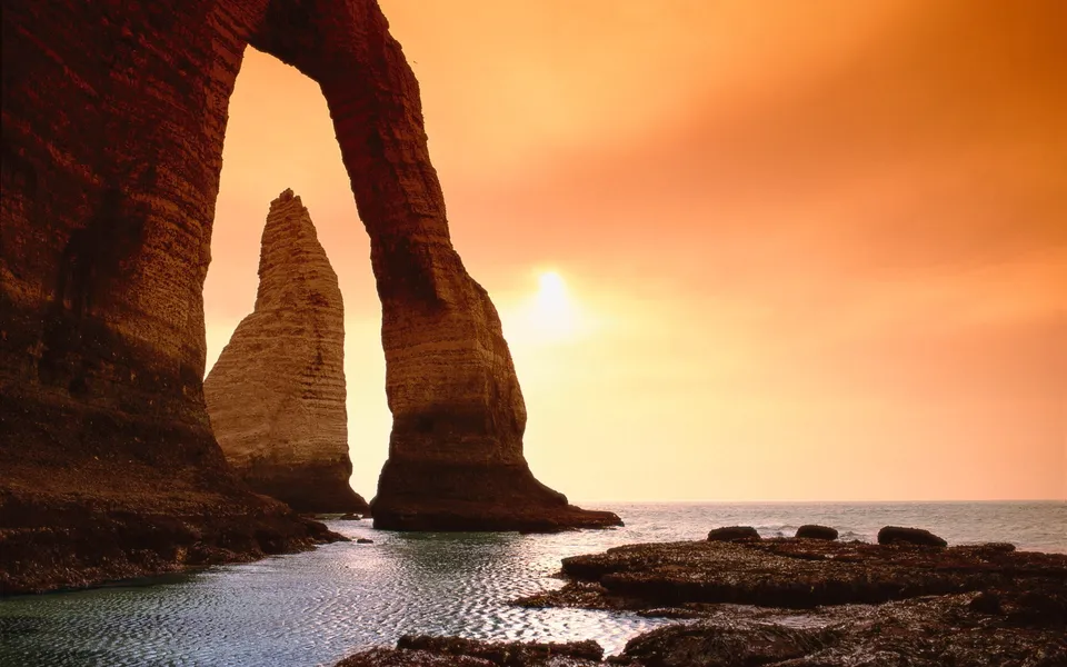 A natural rock arch frames a tall sea stack against a vibrant orange sunset sky over the ocean.