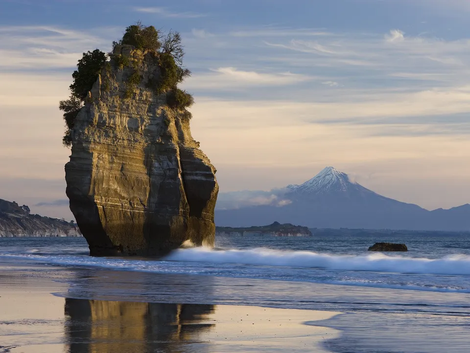 A tall, weathered sea stack stands on a sandy beach with waves crashing at its base. A snow-capped mountain is visible in the distance.