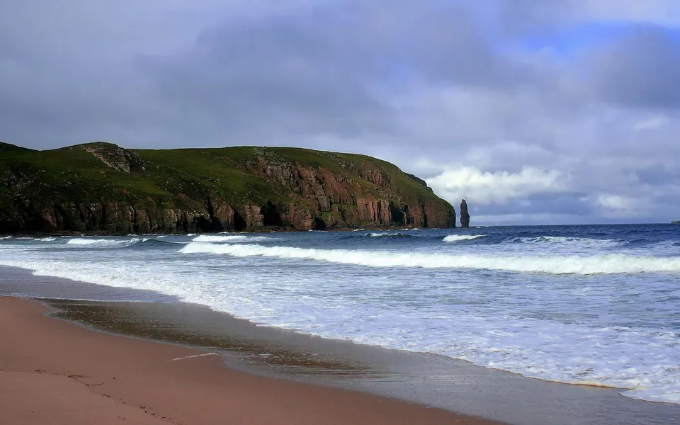 A tall, slender sea stack stands offshore from a green, grassy cliff face with red rock layers.
