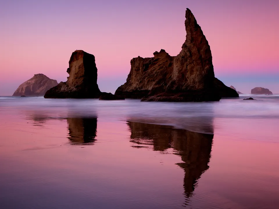 Large rock formations rise from the ocean at sunset, with their reflections visible in the wet sand.