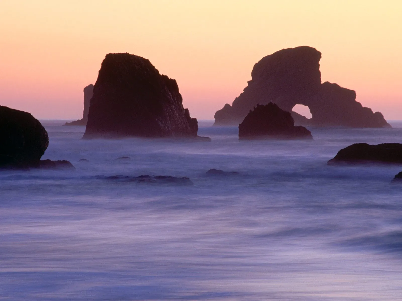 Silhouetted sea stacks and rock formations in the ocean at sunset with soft, misty water.