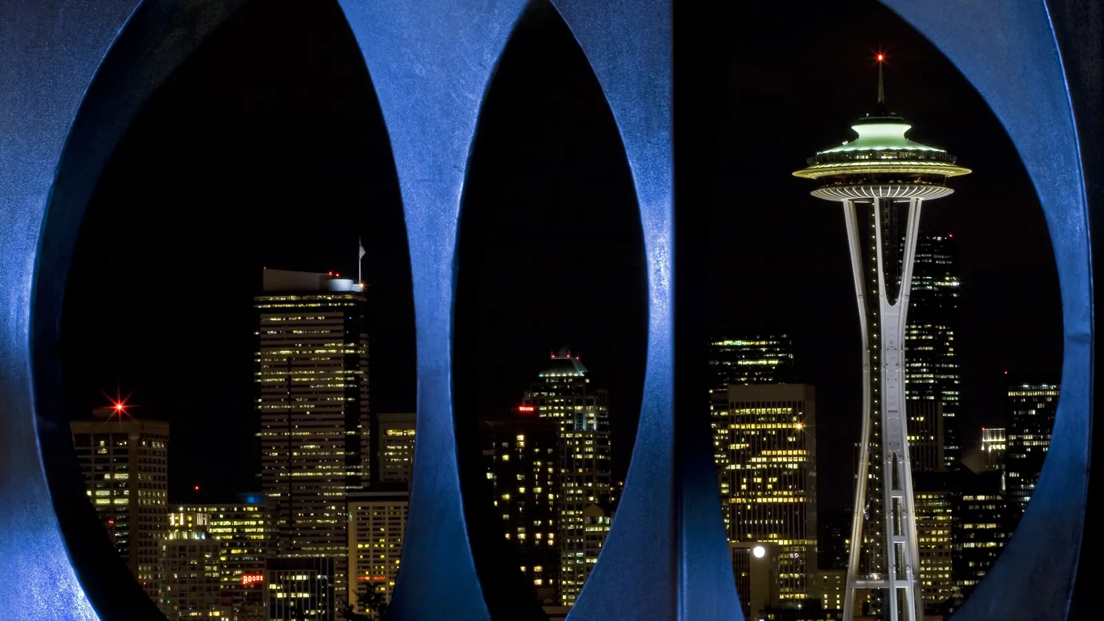 A nighttime view of the Seattle skyline, featuring the Space Needle and illuminated skyscrapers, framed by a dark, abstract sculpture.