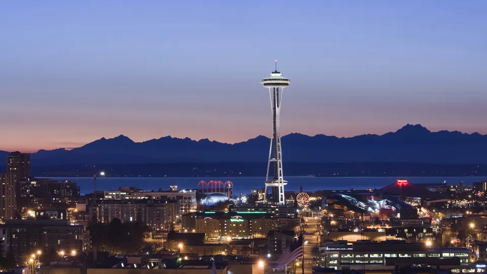 A panoramic view of the Seattle skyline at dusk, featuring the Space Needle and distant mountains.
