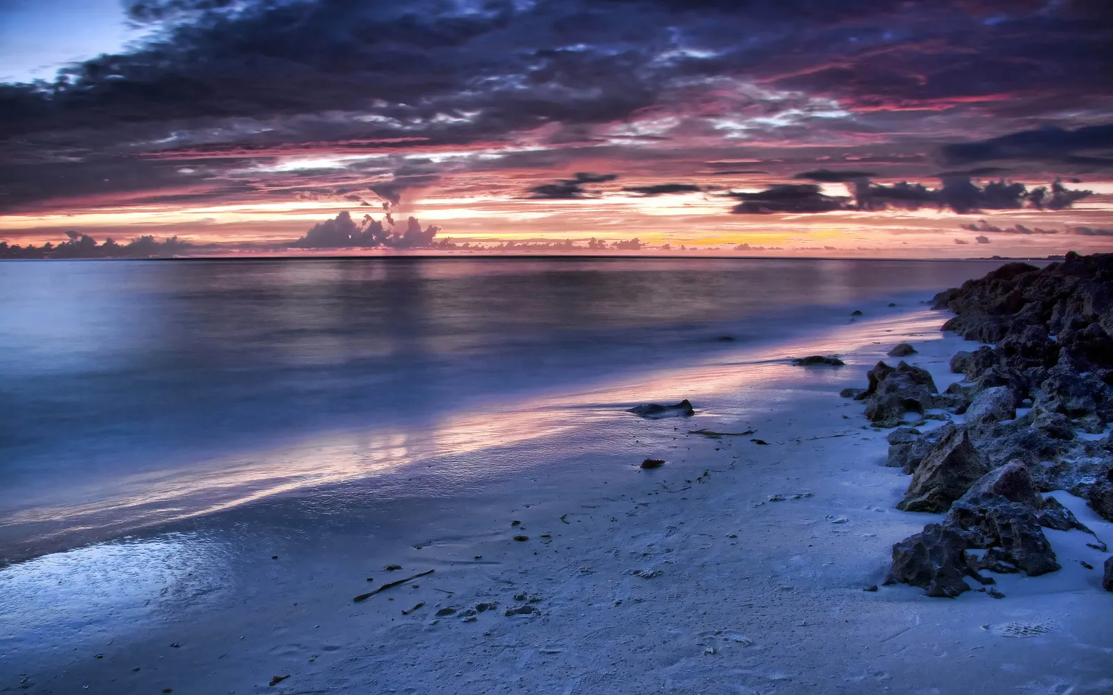Calm ocean reflects a vibrant sunset sky on a sandy shore with rocks.