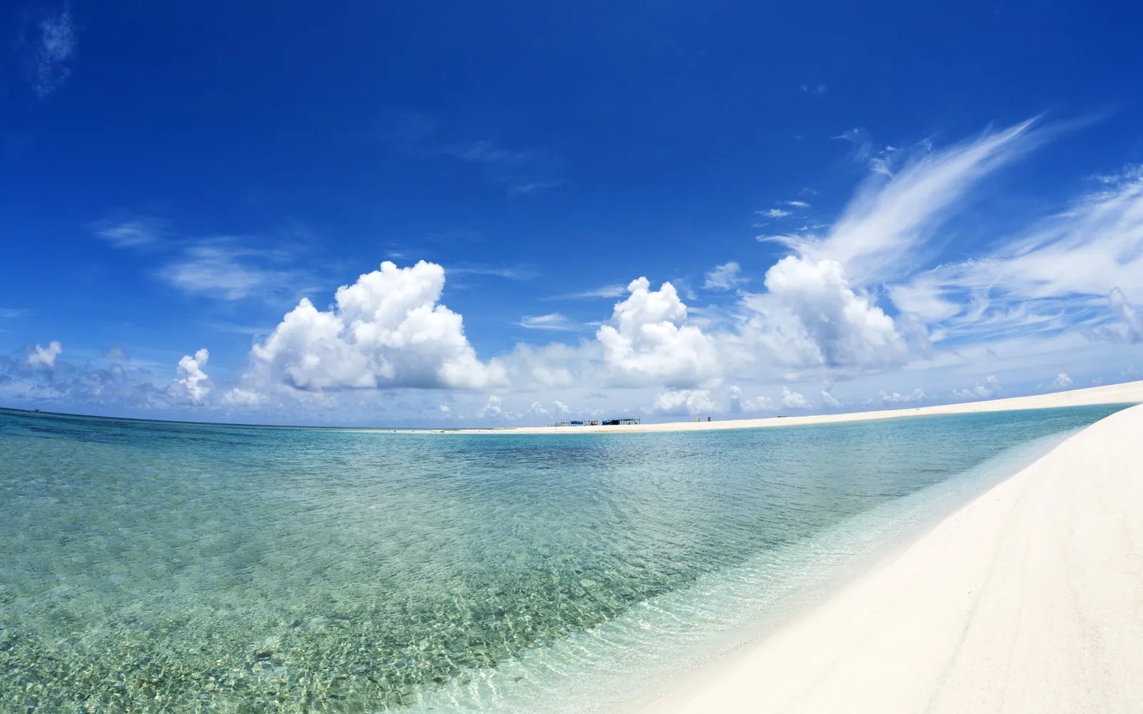 A tranquil tropical beach scene with clear turquoise water lapping on white sand under a blue sky with fluffy clouds.