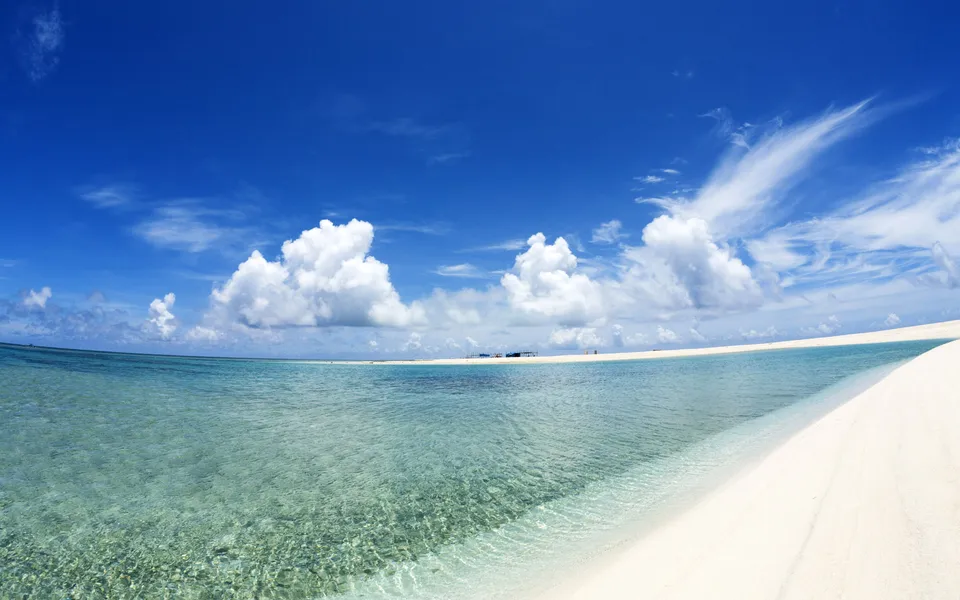 A tranquil tropical beach scene with clear turquoise water lapping on white sand under a blue sky with fluffy clouds.