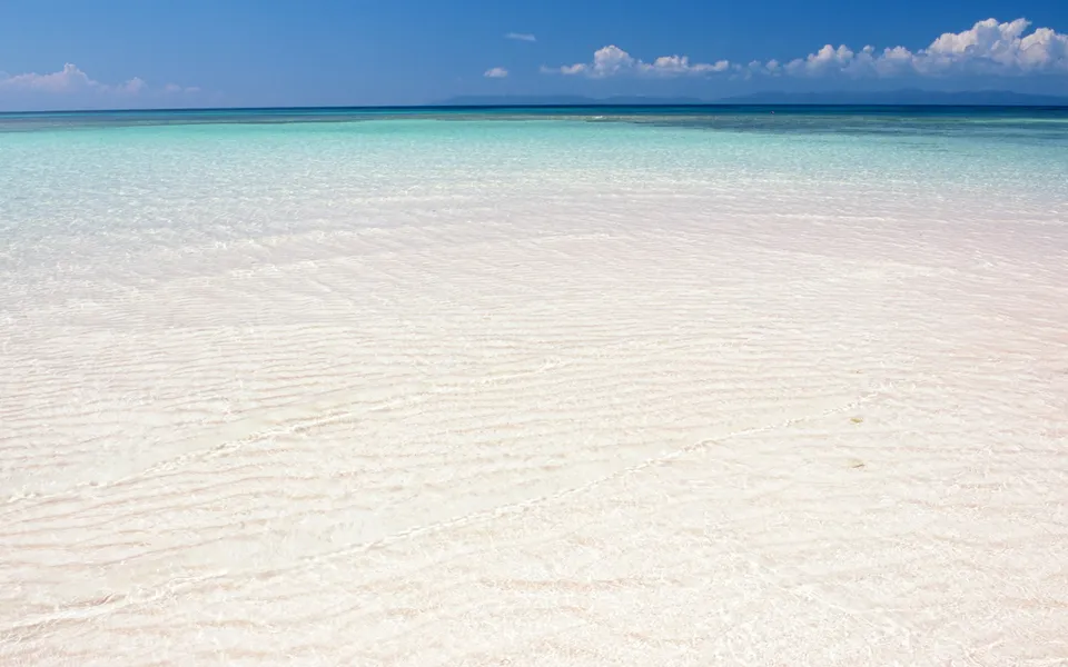 Shallow clear tropical waters showing intricate patterns on the sandy seabed under bright sunlight.