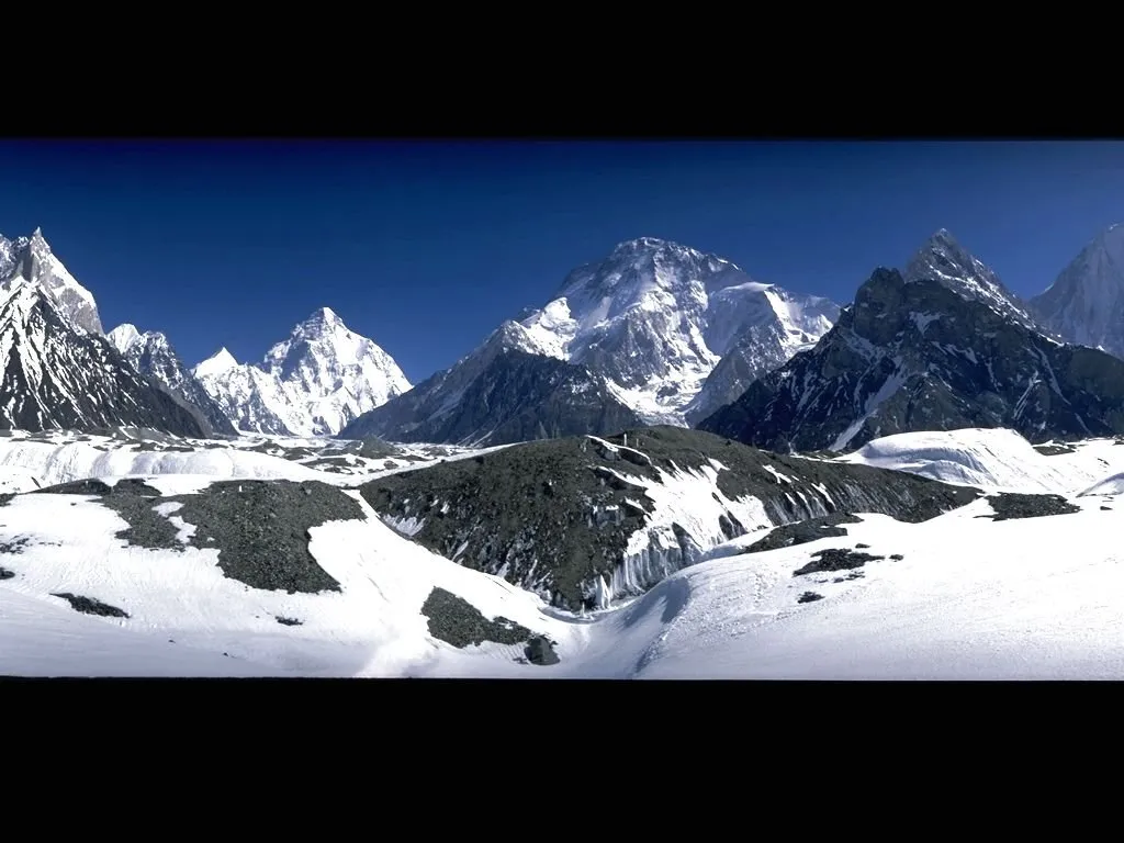 A vast mountain range covered in snow with sharp peaks against a deep blue sky, foreground of snow-dusted rocks and ice.