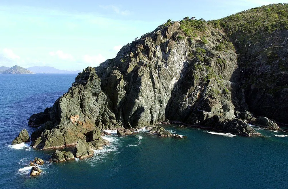 Rugged rocky coastline with sheer cliffs dropping into the dark blue sea under sunlight.
