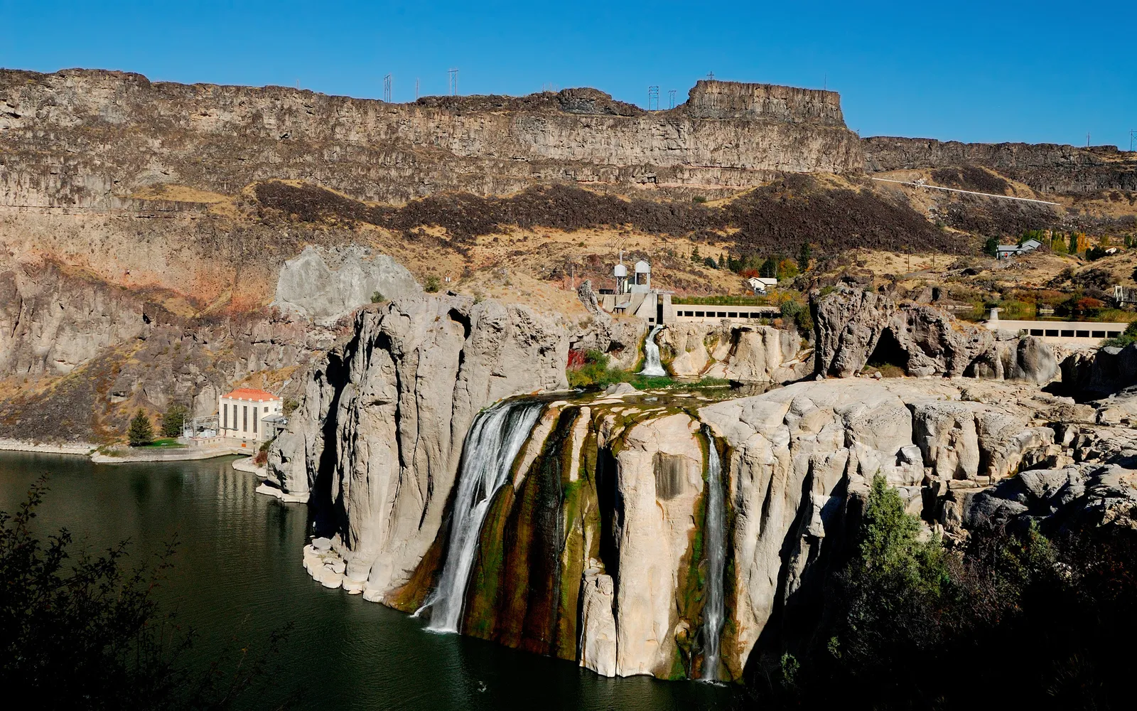Shoshone Falls plunges over a wide, rocky ledge into a dark green river, with cliffs and buildings in the background.