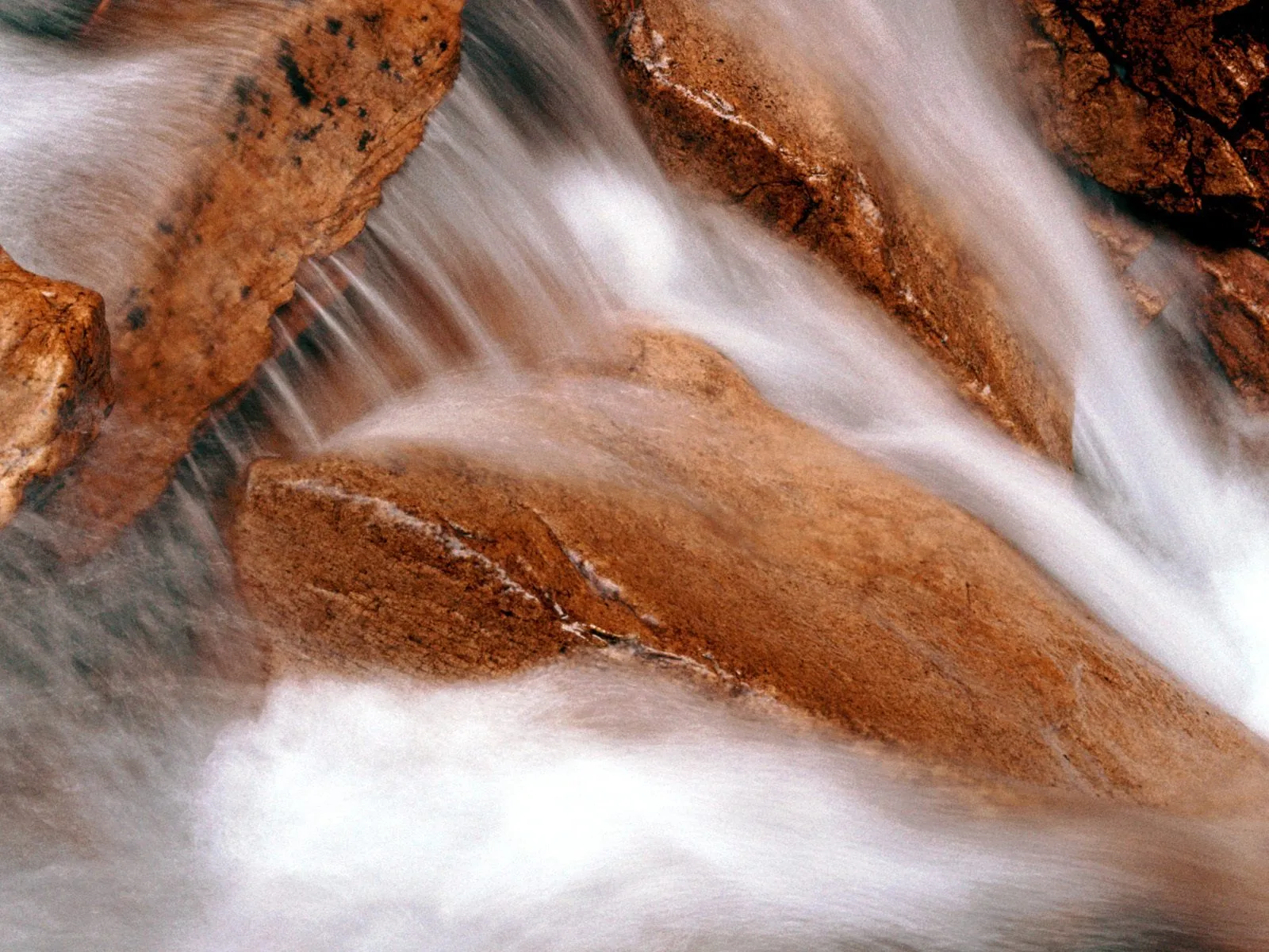 Water flows smoothly over reddish-brown river rocks, creating a silky effect with a long exposure.