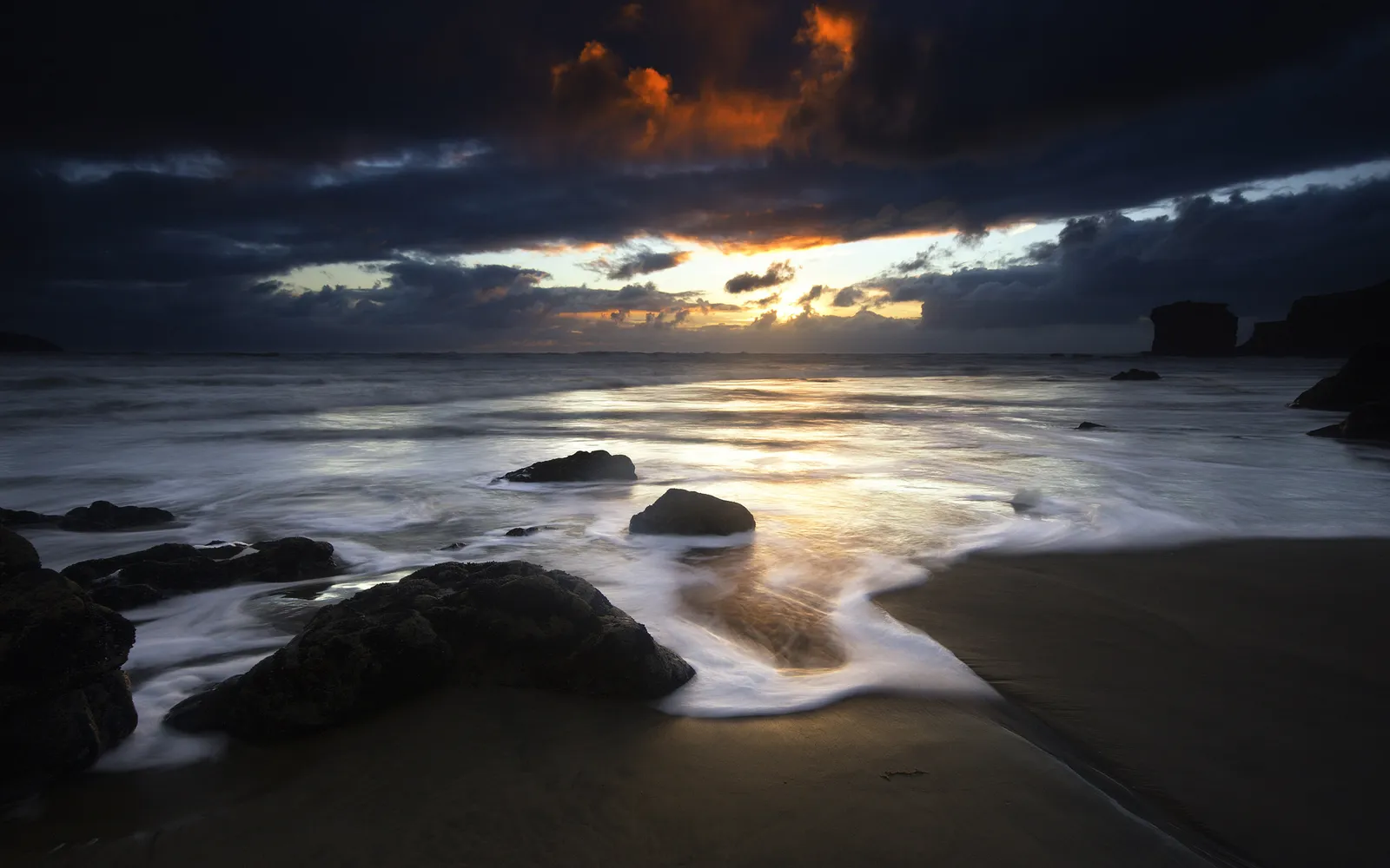 Waves captured in motion as they wash over dark rocks at sunset.