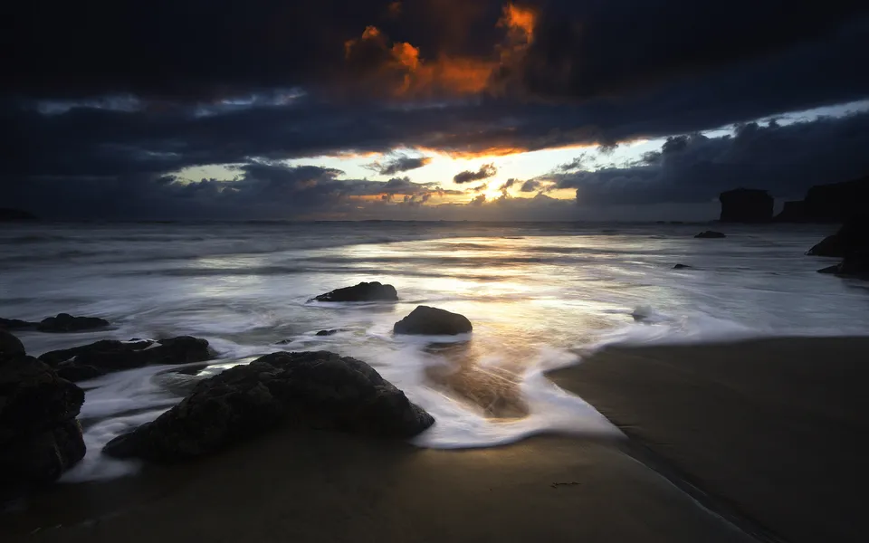 Waves captured in motion as they wash over dark rocks at sunset.