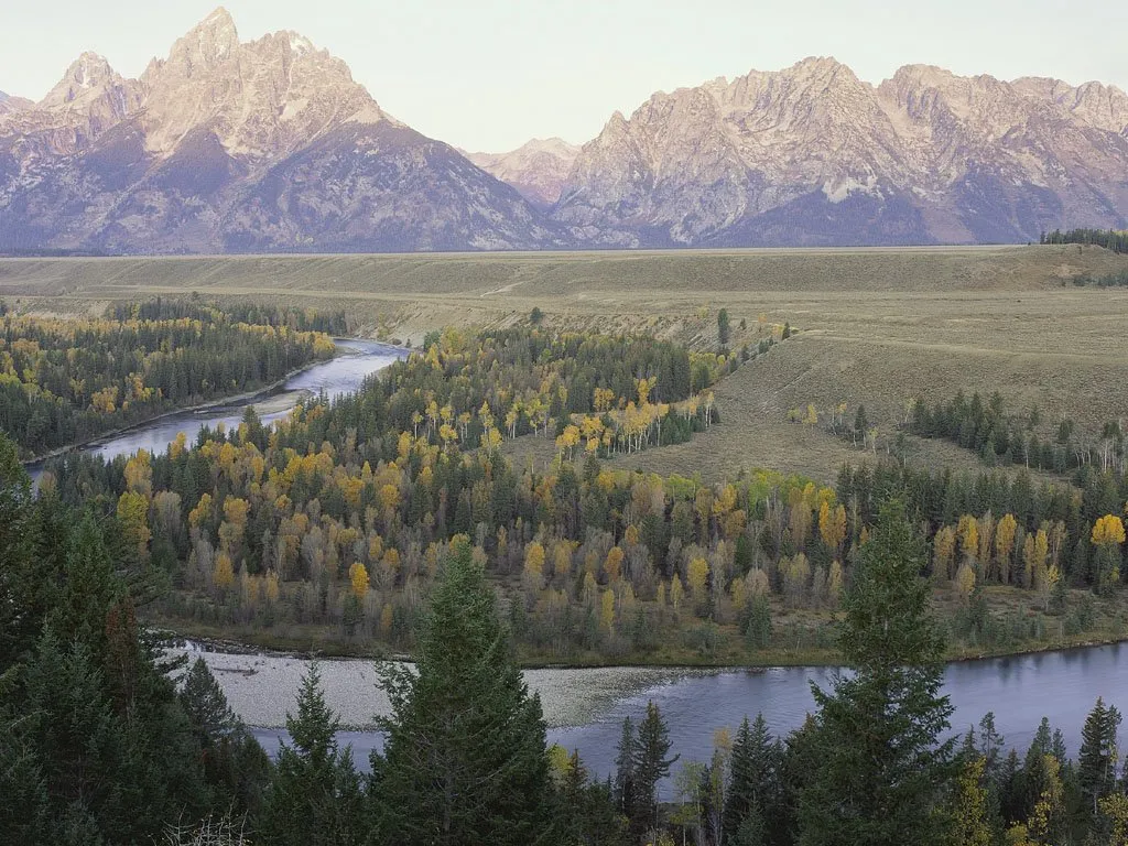 A wide river winds through a valley of colorful autumn trees, with majestic mountains in the background.
