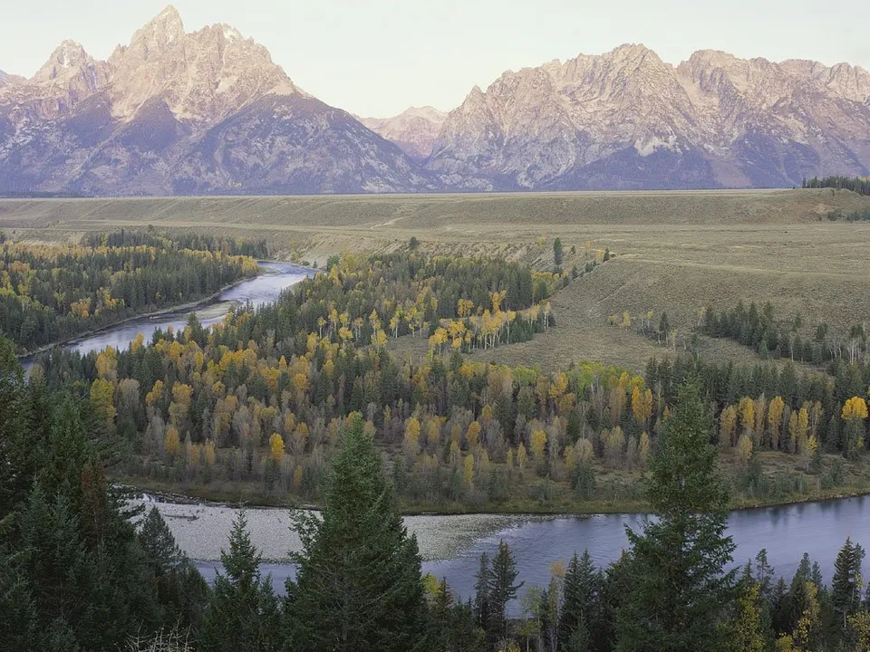 A wide river winds through a valley of colorful autumn trees, with majestic mountains in the background.