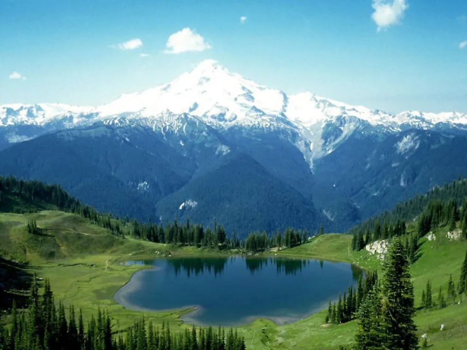 A majestic snow-covered mountain range reflects in a dark blue alpine lake surrounded by green hills and pine trees.
