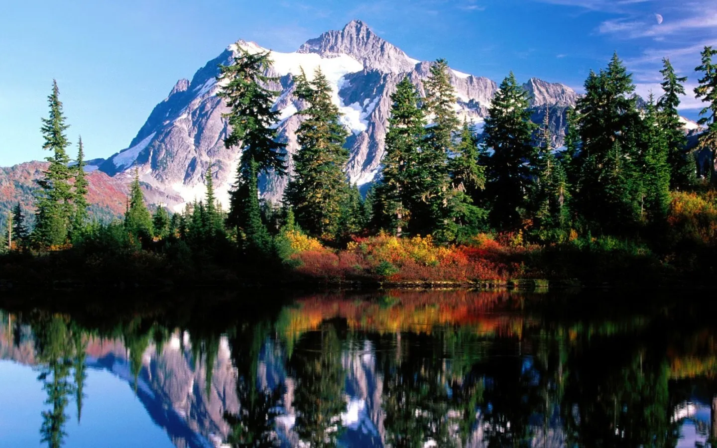A snow-capped mountain reflects in a calm lake surrounded by autumn trees.