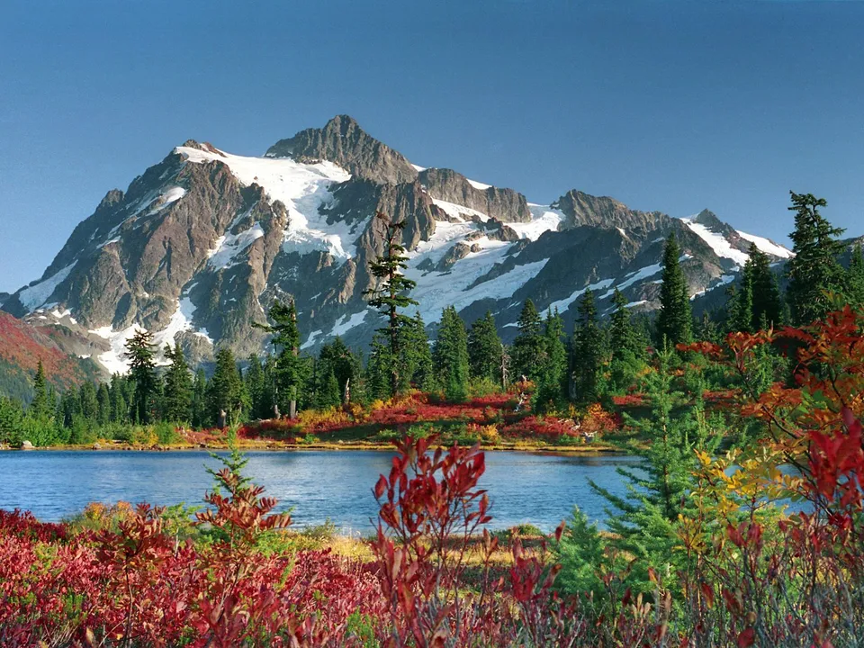 A snow-capped mountain range is reflected in a calm lake surrounded by vibrant red and yellow autumn foliage.