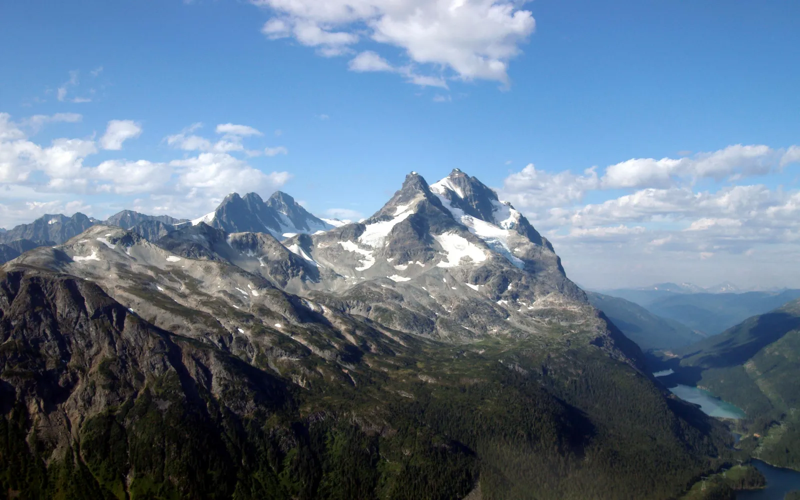 A majestic mountain range with snow-capped peaks and lush green forests below under a cloudy blue sky.