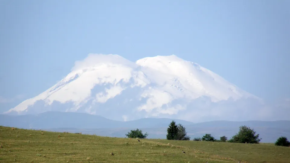 Snow-covered mountain peaks are partially obscured by wispy clouds, with a green meadow in the foreground.