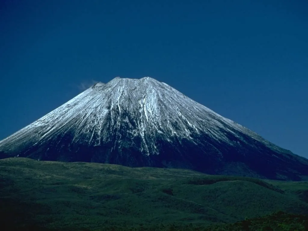 A majestic snow-capped volcano rises against a deep blue sky, with dark green forested foothills in the foreground.
