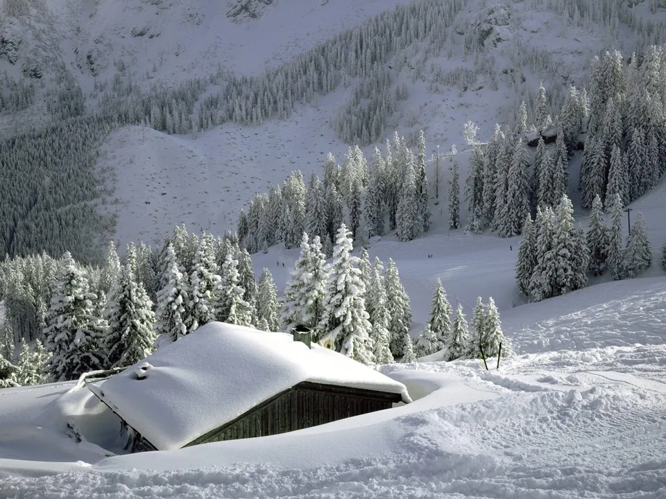A small wooden cabin is almost completely buried in snow, with a backdrop of snow-covered pine trees and mountains.