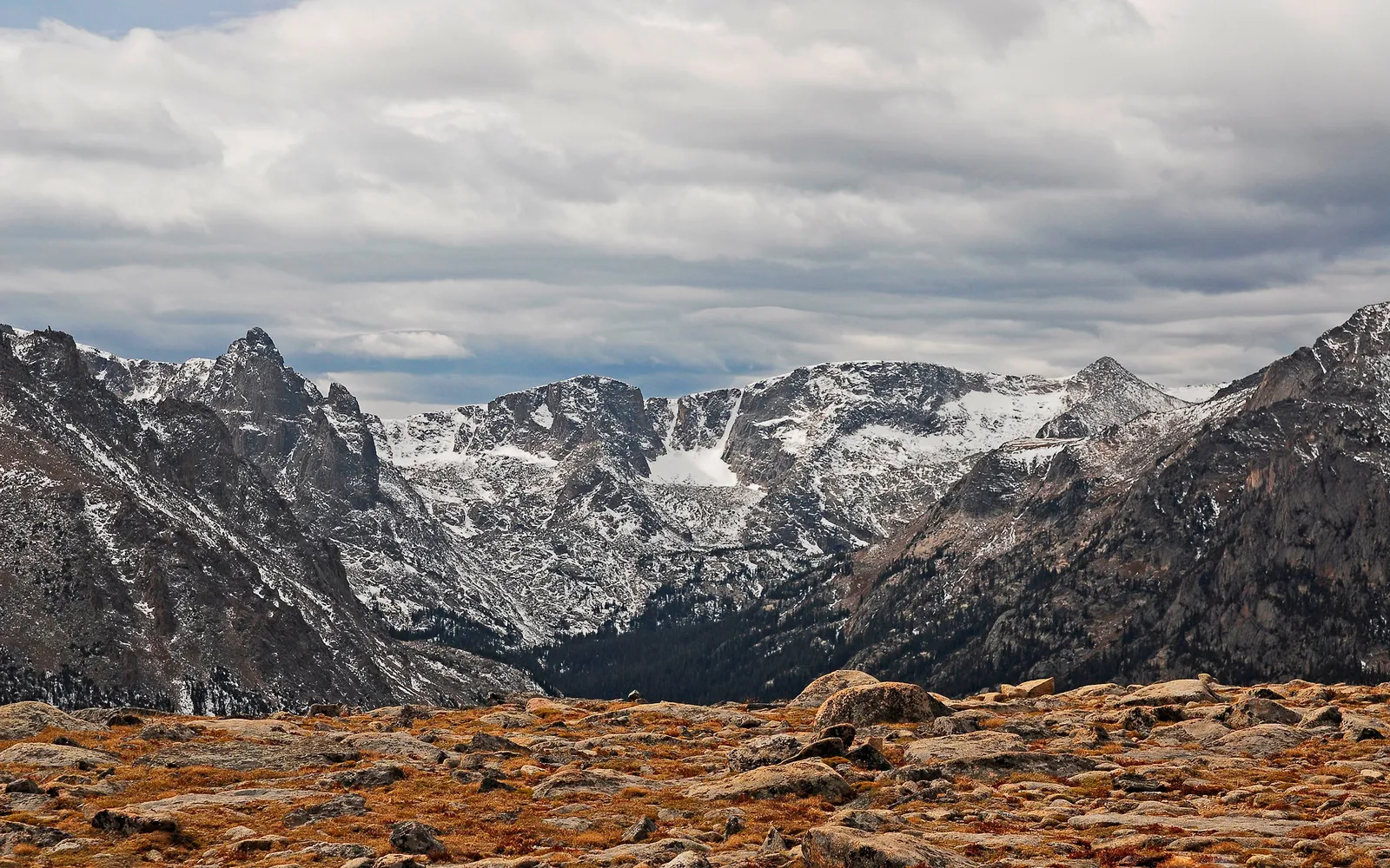 A dramatic mountain range with snow on upper slopes, set against a cloudy sky and rocky, dry grass foreground.