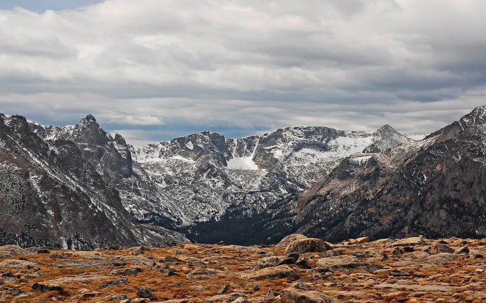 A dramatic mountain range with snow on upper slopes, set against a cloudy sky and rocky, dry grass foreground.