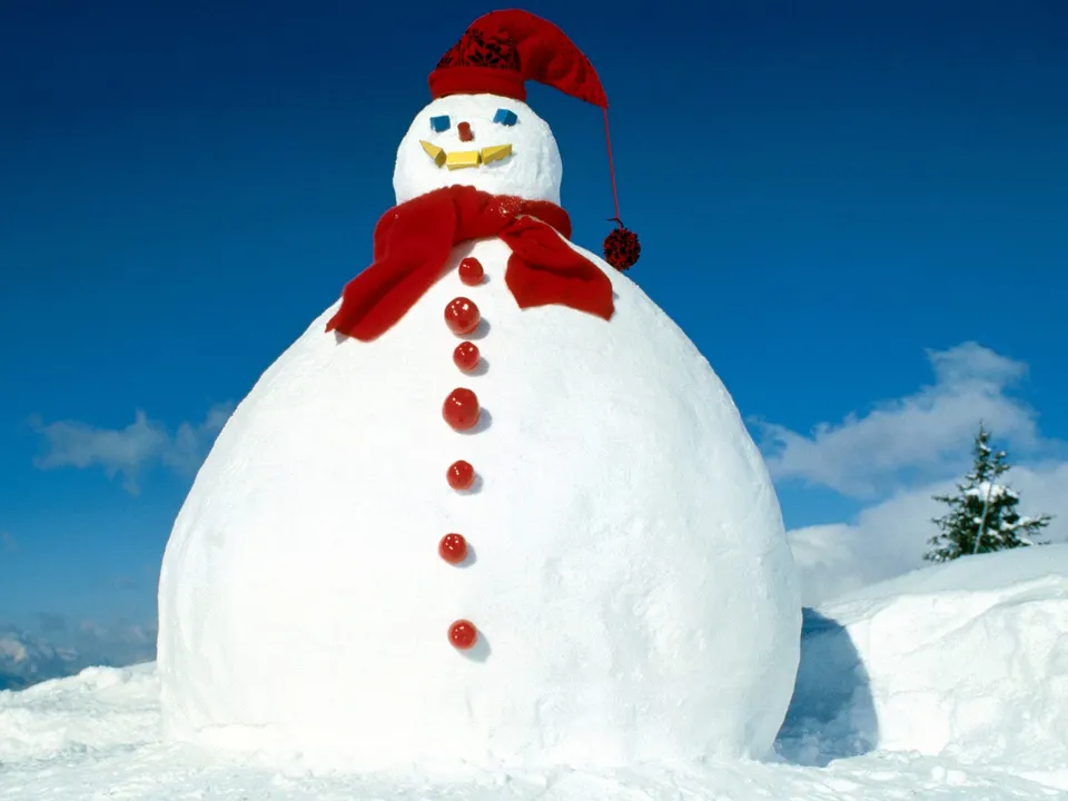 A very large snowman with a red hat and scarf stands in a snowy landscape under a bright blue sky.