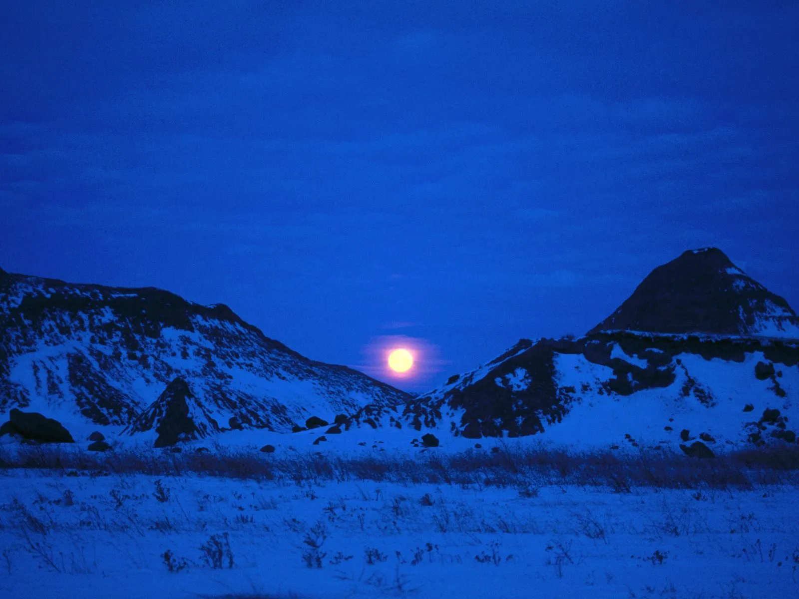 A full moon shines brightly over snow-covered badlands under a deep blue twilight sky.