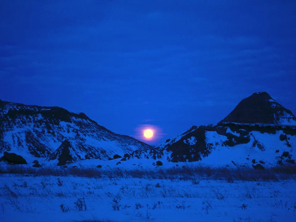 A full moon shines brightly over snow-covered badlands under a deep blue twilight sky.