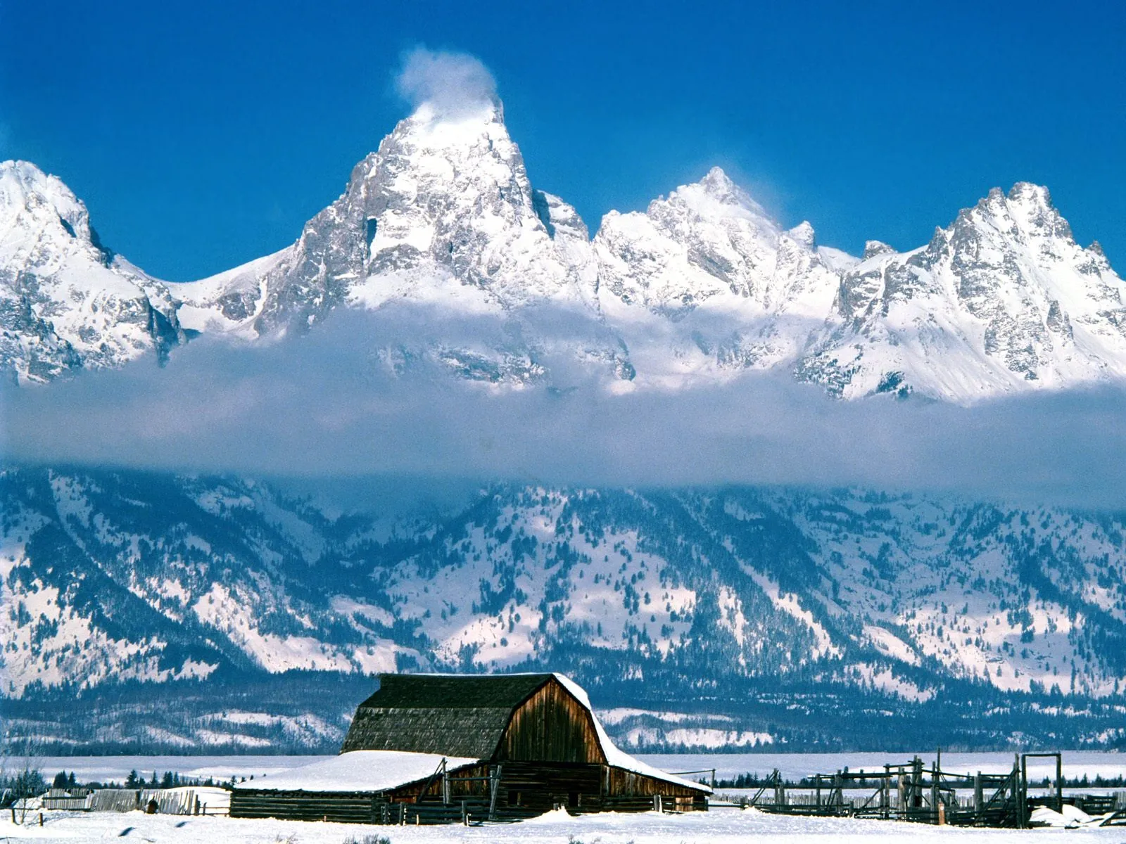 A rustic wooden barn sits in a snowy landscape with majestic, snow-covered mountains rising behind it under a clear blue sky.
