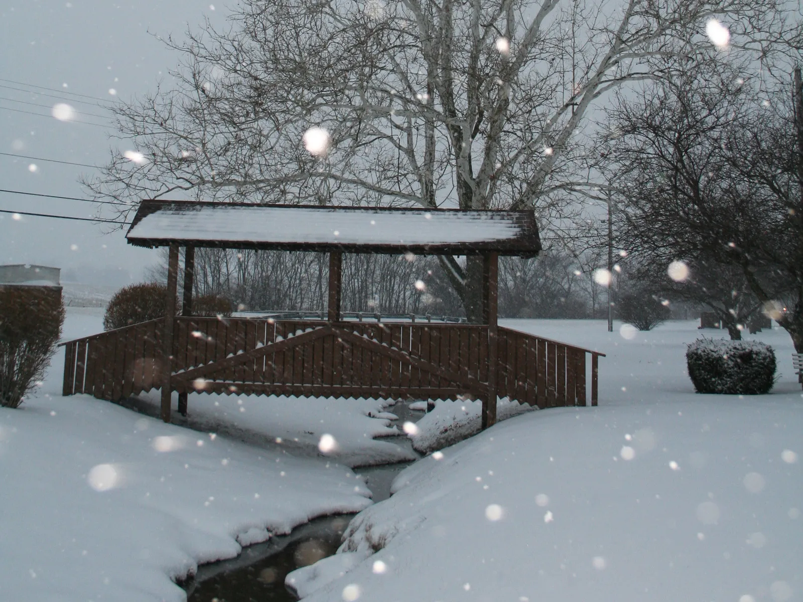 A wooden covered bridge spans a small stream during a snowfall.