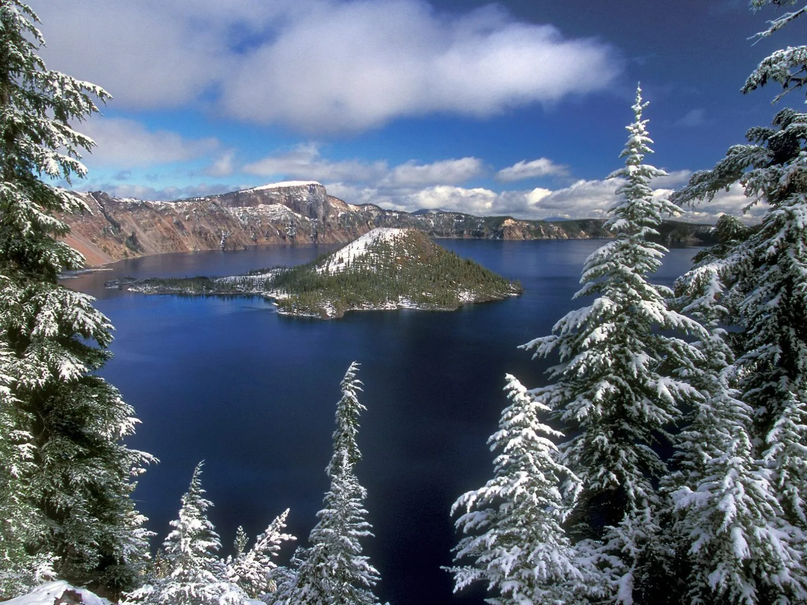 Snow-covered pine trees frame a view of a deep blue lake surrounded by mountains, with a small island in the center.
