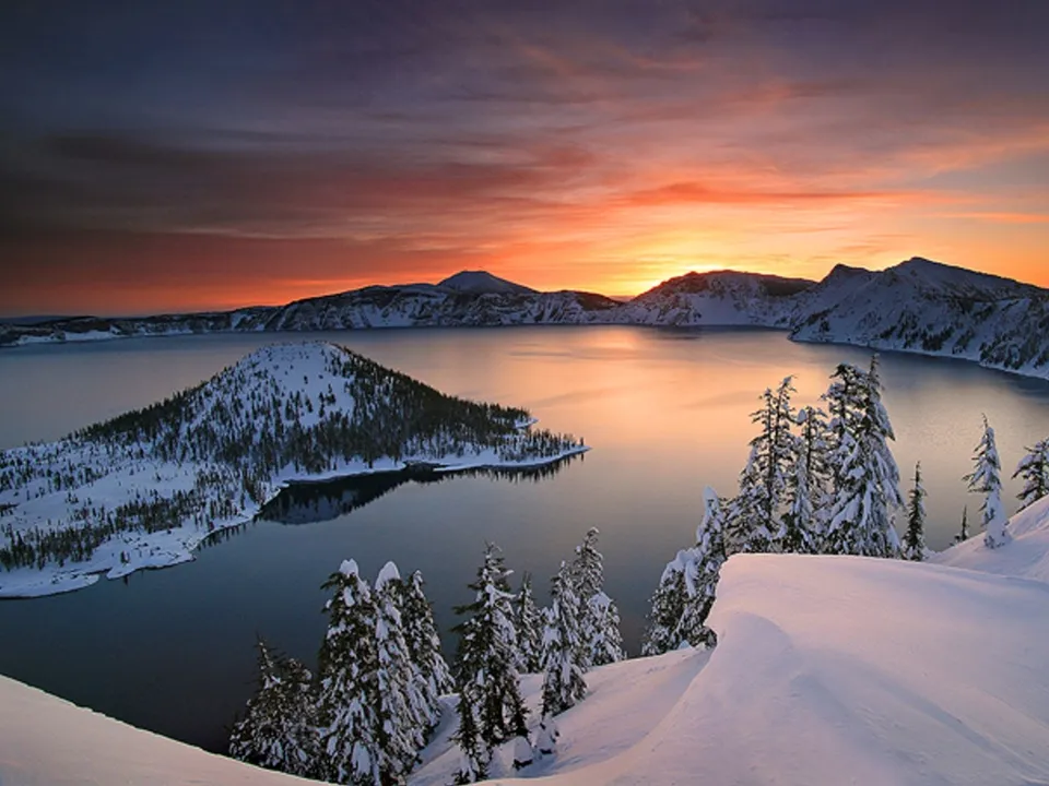 A snow-covered landscape surrounds a vast lake reflecting a vibrant sunset sky with mountains in the background.