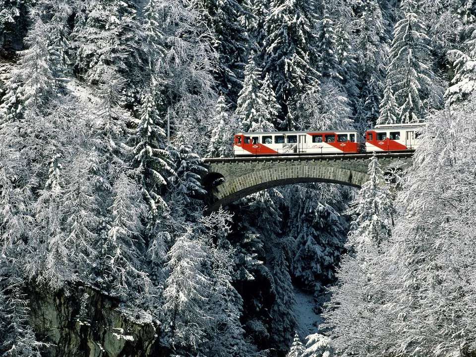 A red and white train travels across a stone bridge in a snow-covered forest.
