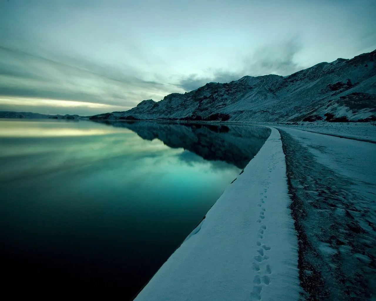 A calm, dark lake reflects the sky and snow-covered mountains under a cloudy sky.
