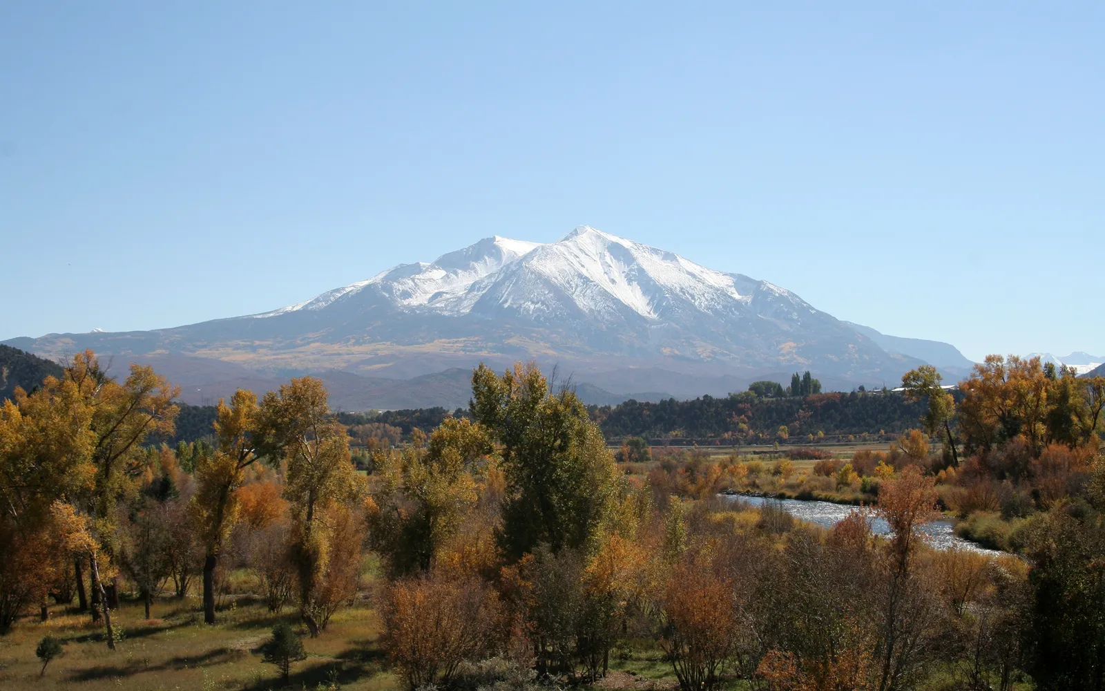 A wide view of a snow-covered mountain range under a clear blue sky, with a valley of autumn trees and a river in the foreground.