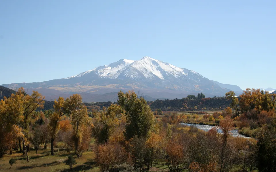 A wide view of a snow-covered mountain range under a clear blue sky, with a valley of autumn trees and a river in the foreground.