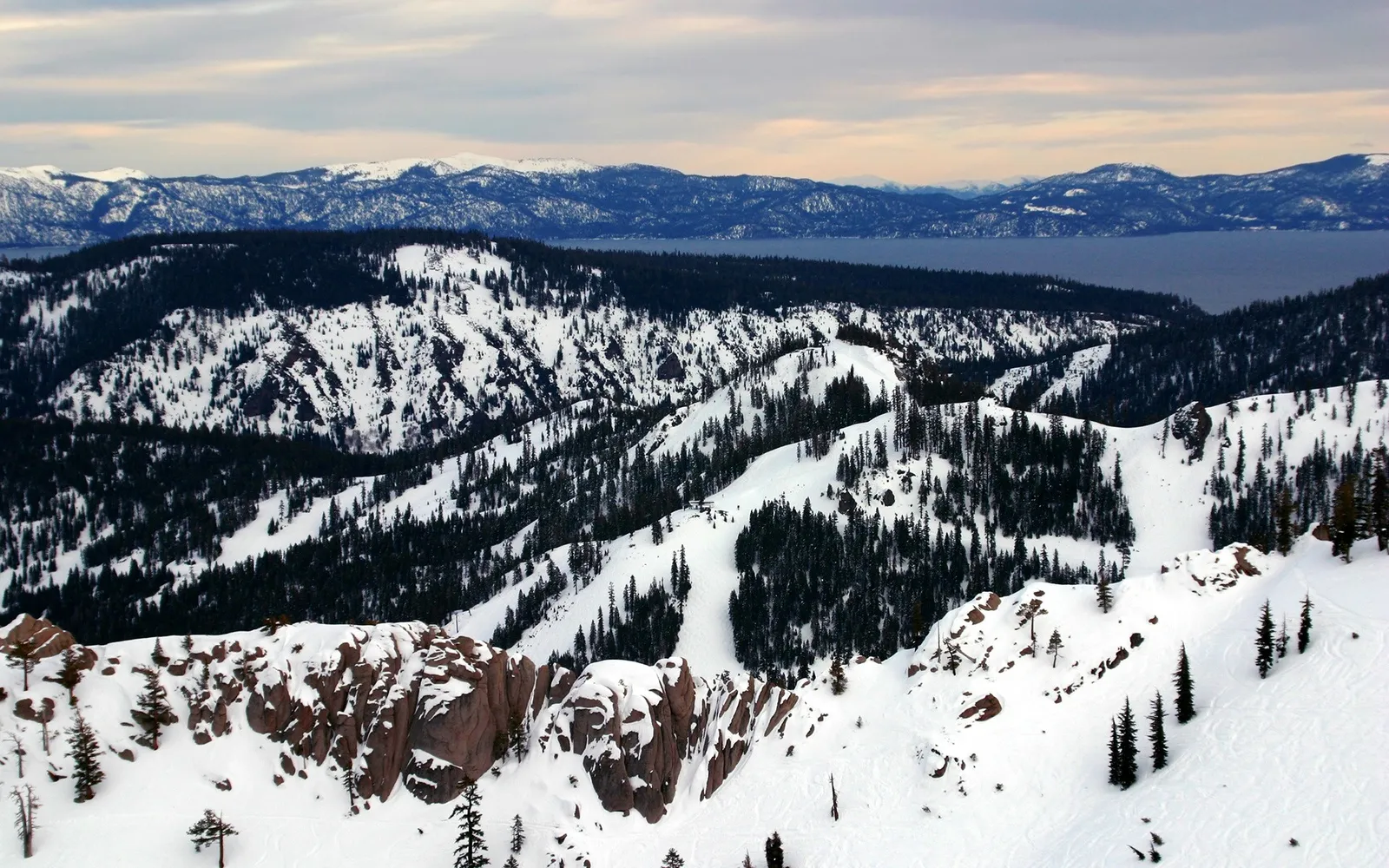 A wide view of snow-covered mountains and pine trees with a large lake in the background under a cloudy sky.